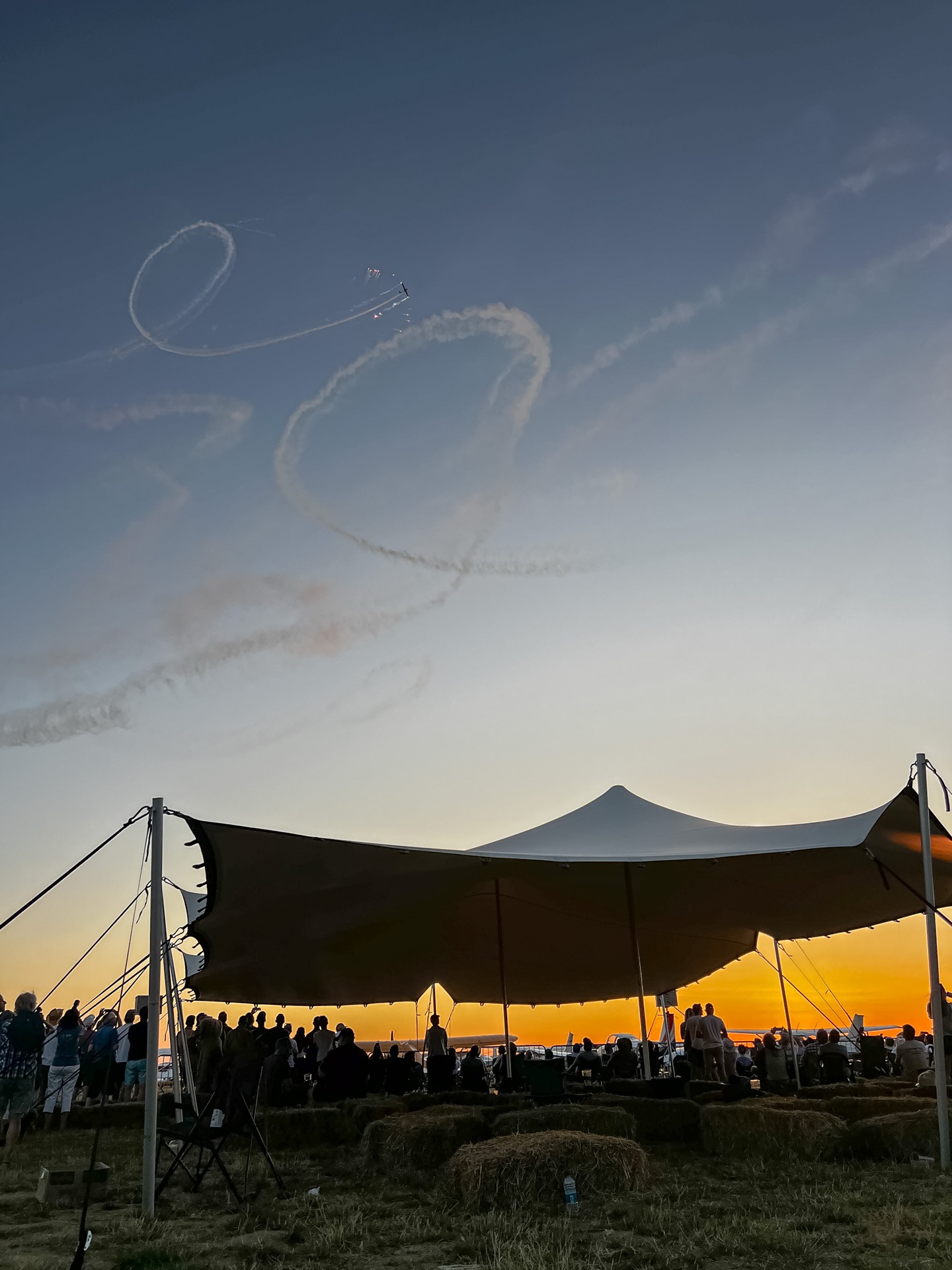 Crowd under a stretch tent at sunset with aerobatic display smoke overhead